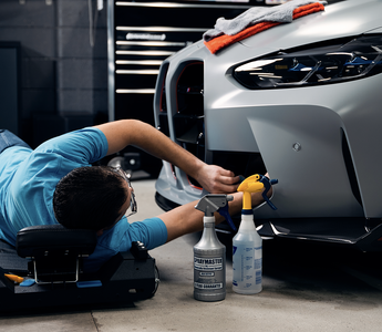 Man in light blue shirt laying down on creeper working on gray car