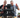 Two guys behind an industrial shop chair on wheels sitting on a table. Both are holding red energy drink cans and smiling