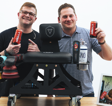 Two guys behind an industrial shop chair on wheels sitting on a table. Both are holding red energy drink cans and smiling